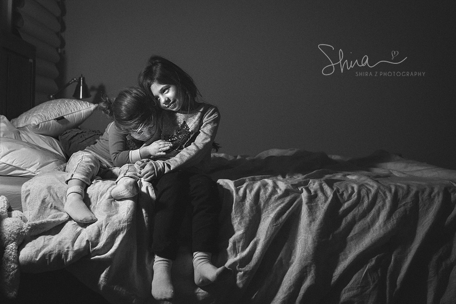 Black and White photo of sisters on a bed leaning on each other photographed by Long Island Family Photographer